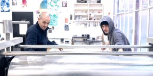 Deux personnes travaillent sur une presse de gravure dans l’atelier de Prépart, un espace lumineux aux murs ornés d’œuvres et de dessins.